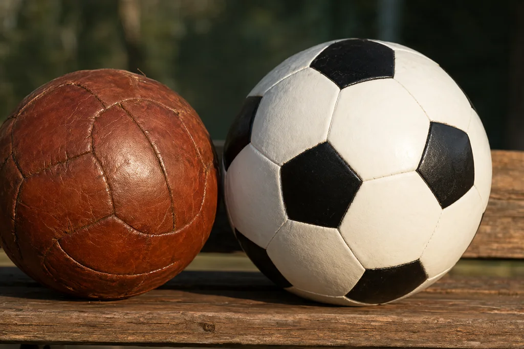 A vintage brown leather round soccer ball with laces beside a modern white round soccer ball with black pentagon panels on grass