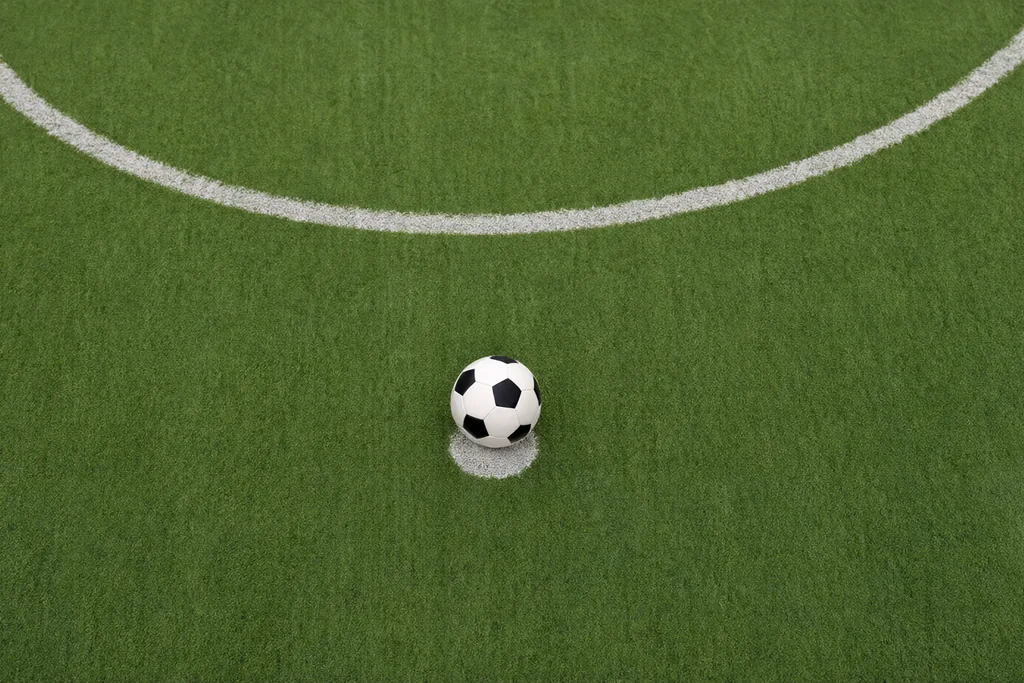 The white painted arc and penalty spot of a green soccer pitch seen from above with a classic round soccer ball on the spot